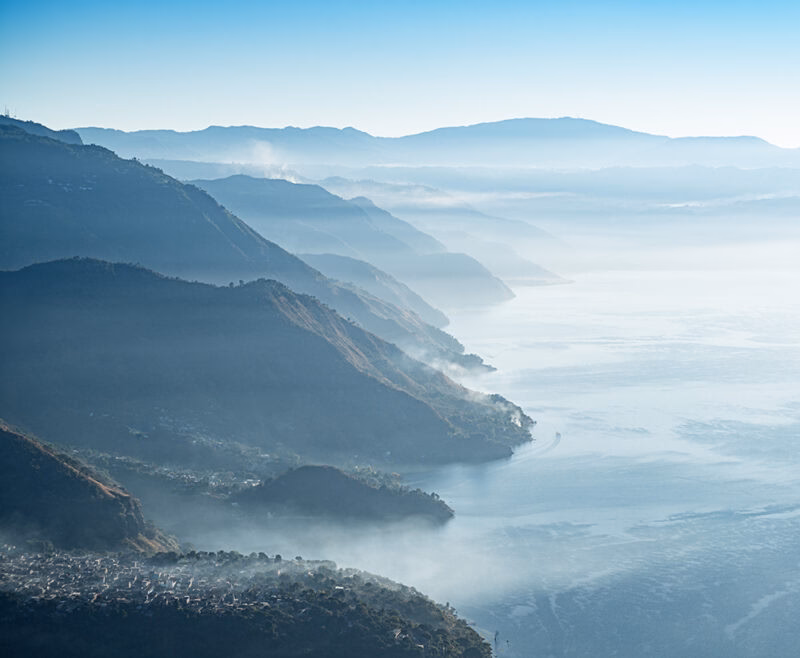 Elevated misty and foggy landscape view of a large blue lake surrounded by steep, layered mountains under a pale blue sky.