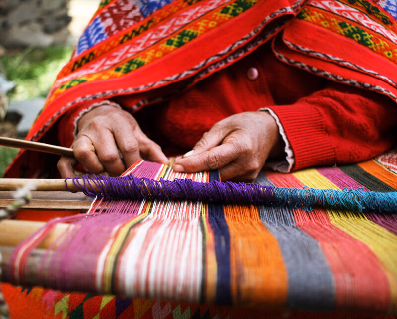 A close-up shows hands weaving a colorful, patterned textile on a wooden loom, with a red cloak draped over the person, ideal for luxury Machu Picchu trips.