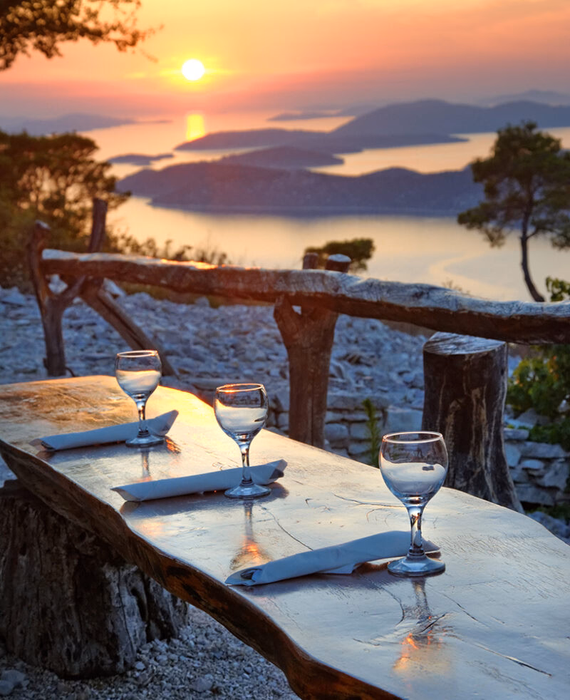 Wine glasses on a wooden table overlooking a sunset and island archipelago on luxury Croatia trips.