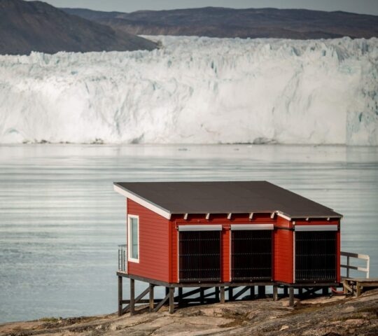 A red wooden cabin on stilts near water and a giant glacier during luxury Polar vacations.