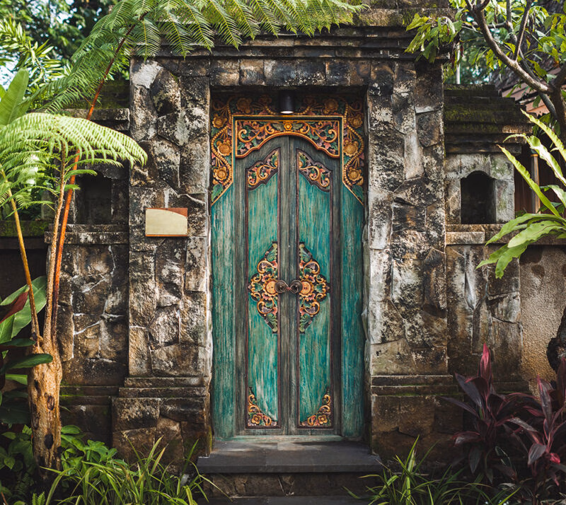 A richly carved, teal and gold double wooden door set in a traditional stone gateway, surrounded by foliage for luxury Indonesia vacations.
