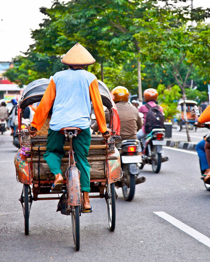 A man in a conical hat and bright shirt rides a becak (cycle rickshaw) down a street. Traffic and trees are in the background, offering luxury Indonesia tours.