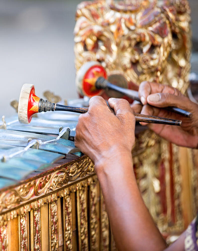 Close-up of hands playing a gamelan instrument with mallets. The ornate, carved wooden frame is gold, highlighting luxury Indonesia trips.