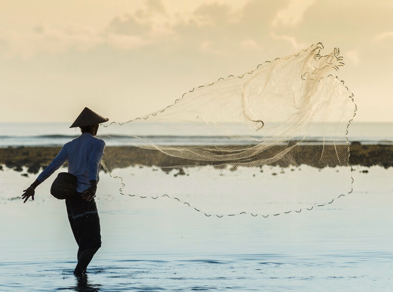 A fisherman in a conical hat casting a large net into the water near the shore at sunset, a scene for luxury Indonesia tours.