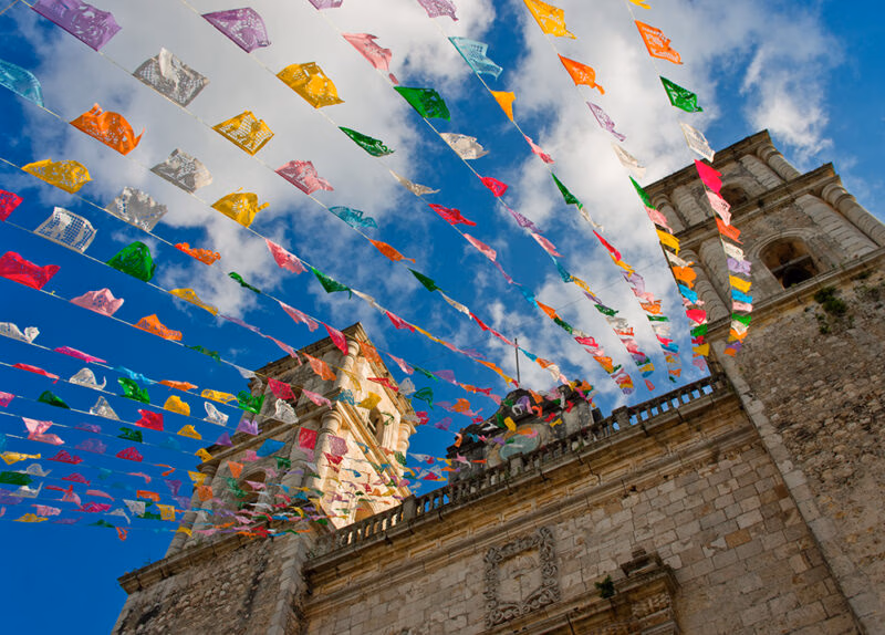 Colorful papel picado banners hang above the stone towers of a colonial church against a blue sky, for luxury Mexico holidays.
