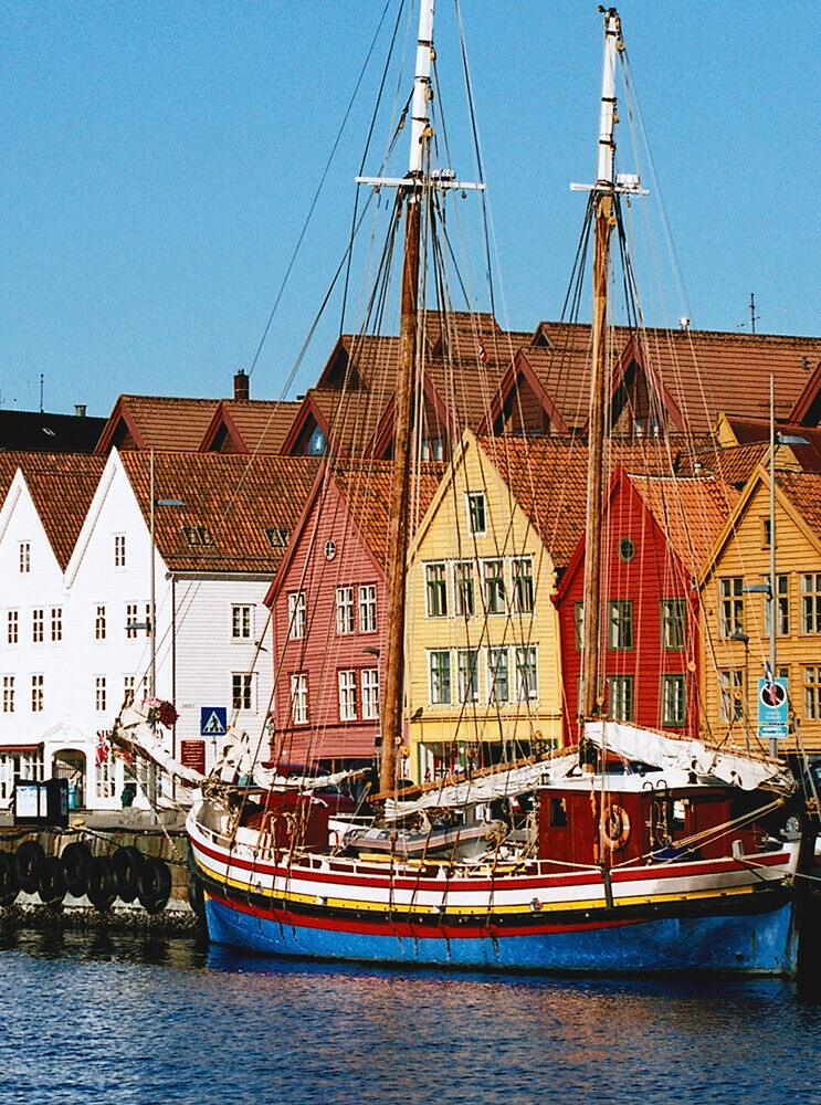 A traditional red, blue, and yellow sailboat docked near historic Bryggen houses in Bergen, Norway, great for luxury Norway tours.