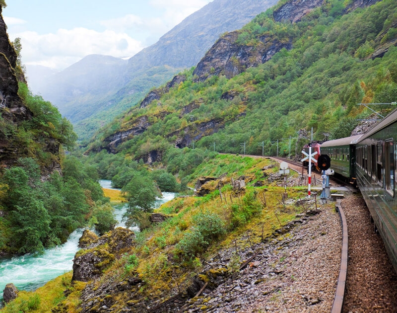 A green train curves along a railway track through a lush, steep valley with a vibrant river, part of luxury Norway tours.
