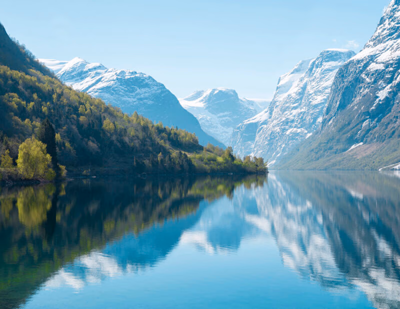 A calm fjord reflects snow-capped mountains and green slopes under a bright blue sky, perfect for luxury Norway trips.