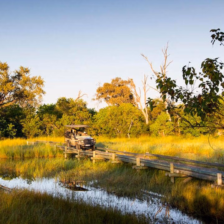 A white safari vehicle driving across a rustic wooden bridge over a small stream in a grassy field.