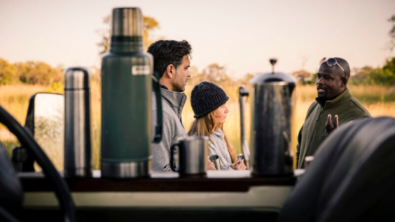 Tourists and a guide talking outdoors with coffee equipment in the foreground.