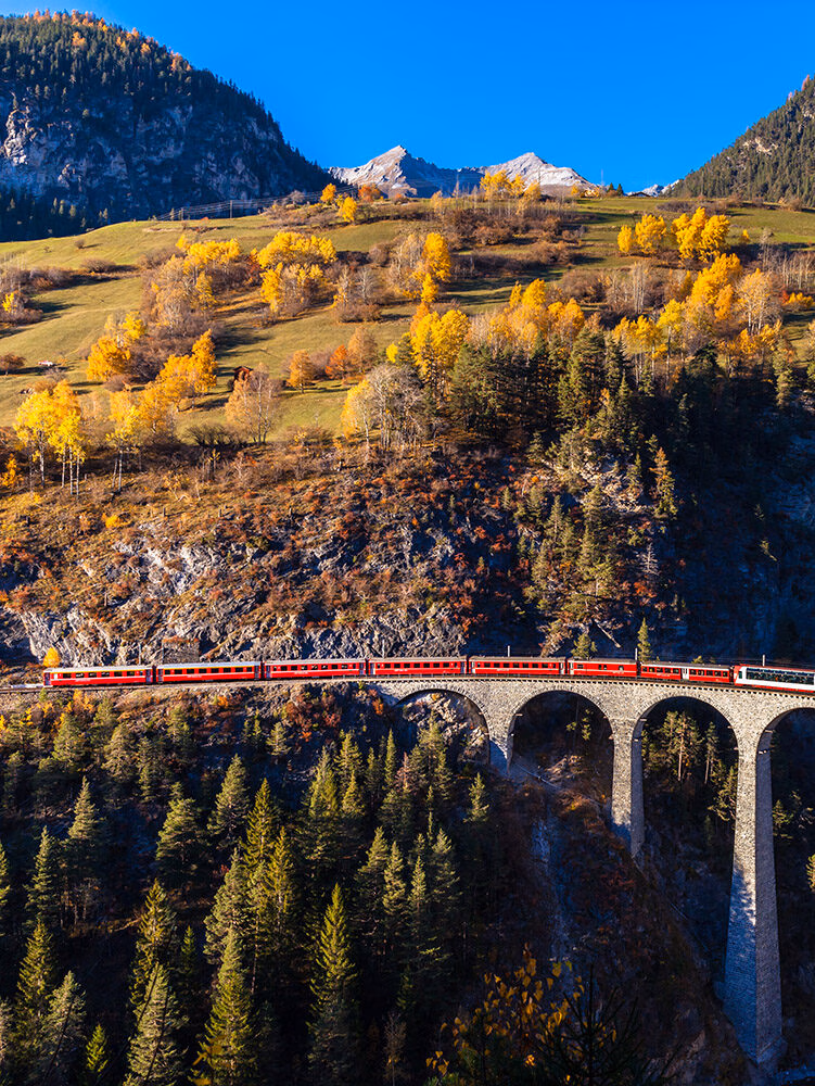 Iconic red train crossing a high arched stone bridge surrounded by golden autumn trees on luxury Switzerland trips.
