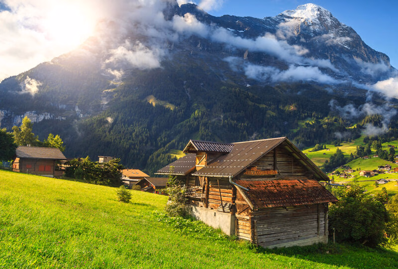 Traditional wooden mountain hut in a sunlit green meadow with snowy peaks for luxury Switzerland vacations.