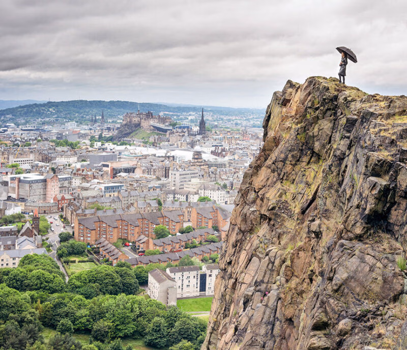 A person stands on the edge of a sheer cliff face overlooking the historic cityscape of Edinburgh and Edinburgh Castle. A highlight of luxury Scotland trips.