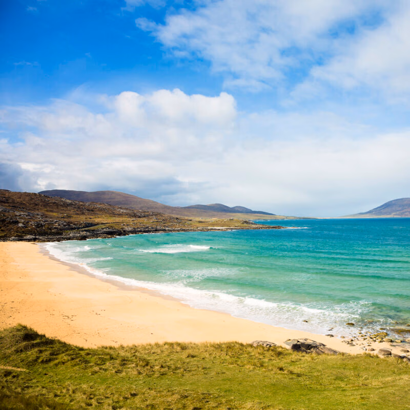A wide, sandy beach with turquoise water and waves breaking onto the shore, surrounded by grassy hills and blue sky. Ideal for luxury Scotland trips.