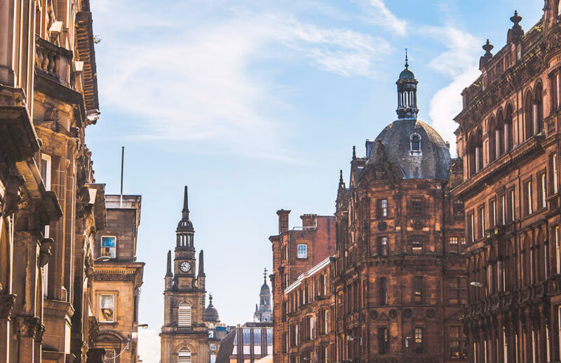 Ornate, historic, red sandstone buildings line a street in Glasgow, with a clock tower and dome against a blue sky. Discover this on luxury Scotland vacations.