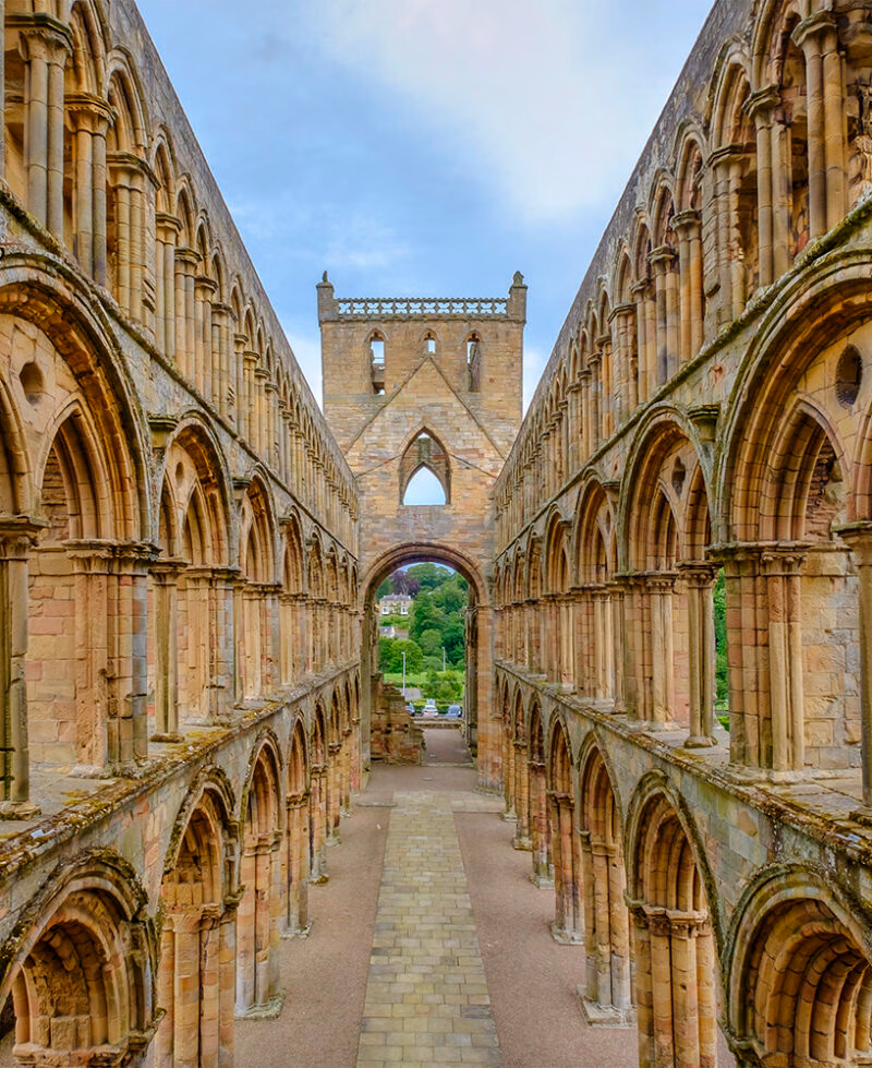 A long corridor view between two rows of symmetrical stone arches inside the ruins of Jedburgh Abbey. A great stop on luxury Scotland trips.