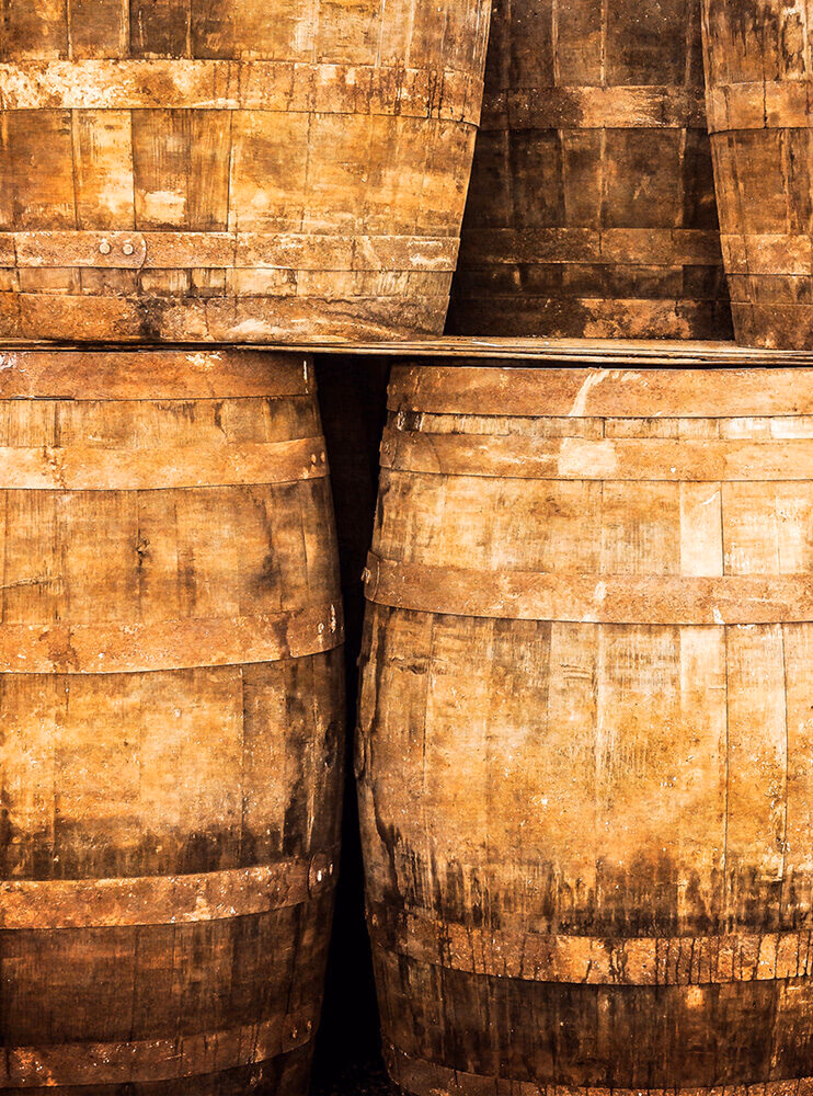 A close-up image of rough, aged wooden barrels stacked tightly together in a warm, brown-orange tone. Explore this aspect of luxury Scotland vacations.