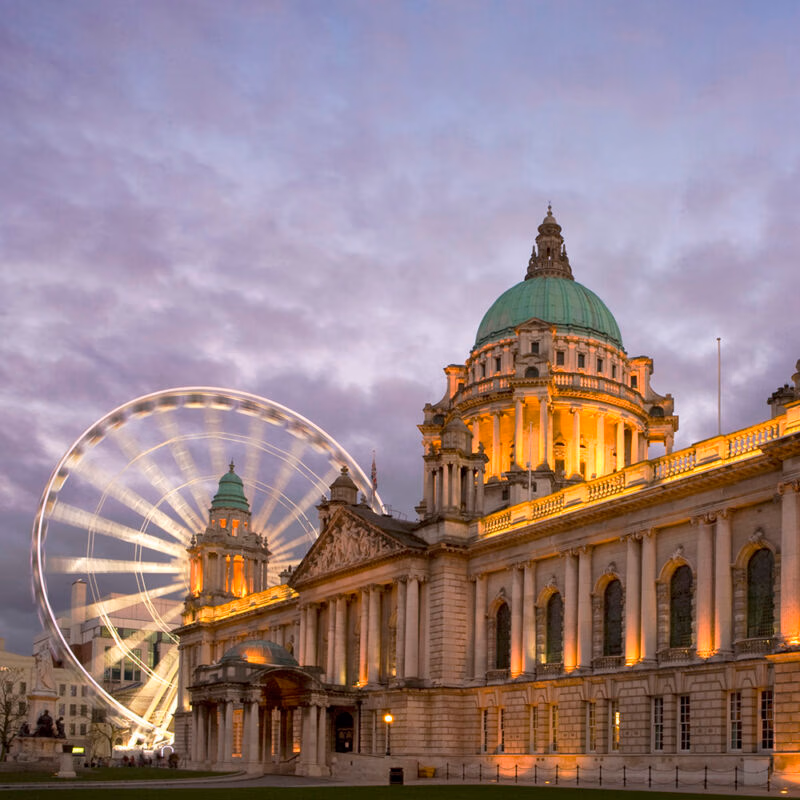 Belfast City Hall, a grand building with an illuminated dome, shown at dusk with a blurred, illuminated Ferris wheel on the left. See landmarks on luxury Ireland trips.