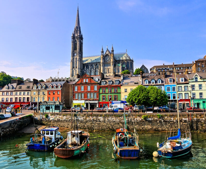 The waterfront of Cobh, Ireland, featuring colorful historic buildings, docked boats, and St. Colman's Cathedral in the background. Consider luxury Ireland holidays for your next trip.