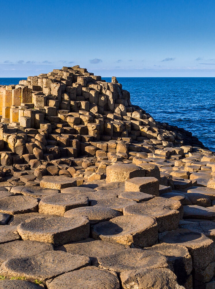 A vertical photo of the Giant's Causeway in Northern Ireland, showing the hexagonal basalt columns meeting the clear blue ocean and sky. Plan your visit on luxury Ireland vacations.