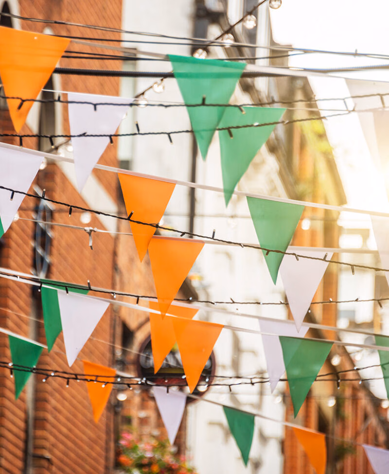 Irish tricolor bunting (orange, white, and green) and fairy lights strung across a street, against a blurred brick building background, offering inspiration for luxury Ireland vacations.