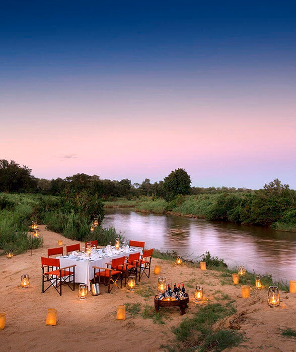 An outdoor dining setup with a long table, red chairs, and many glowing lanterns by a river at dusk.