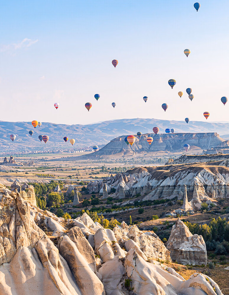 Dozens of colorful hot air balloons float over the dramatic, rocky landscape of Cappadocia, Turkey, featuring unique "fairy chimney" formations.