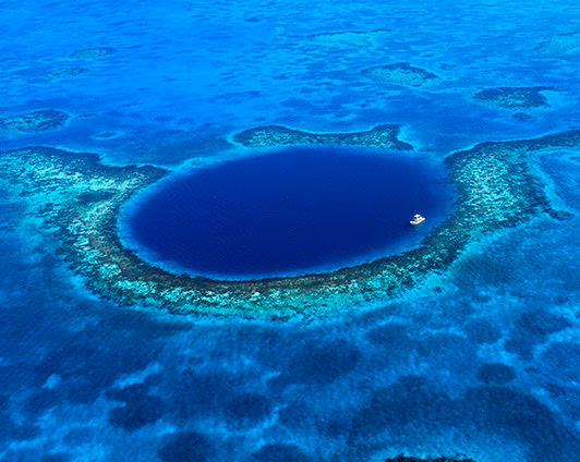 An aerial shot of a large, circular dark blue underwater sinkhole surrounded by turquoise coral reef and a small white boat.