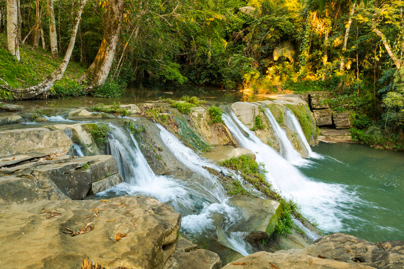 A scenic tropical waterfall flowing into a river within a lush green forest on a sunny day.