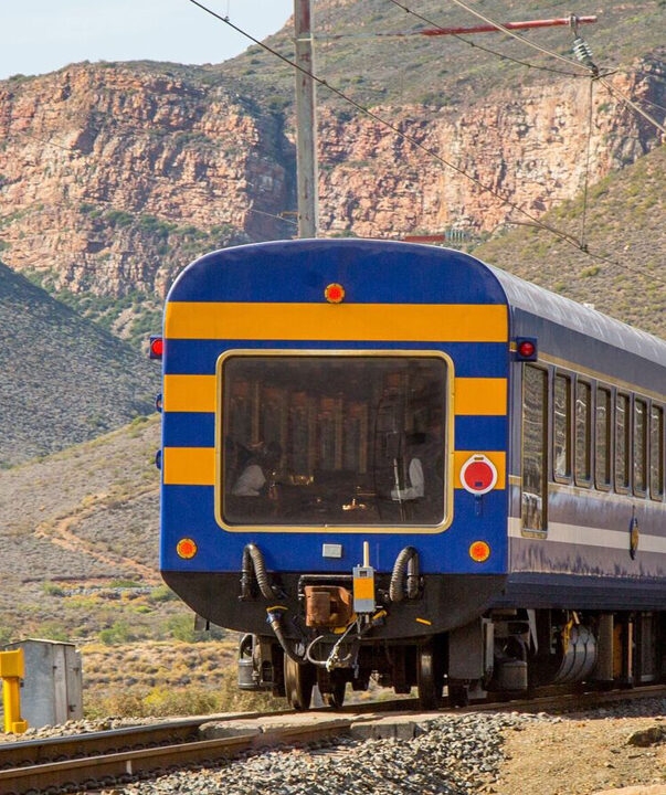 A blue and yellow train moves through a rocky mountain pass on luxury South Africa rail travel.