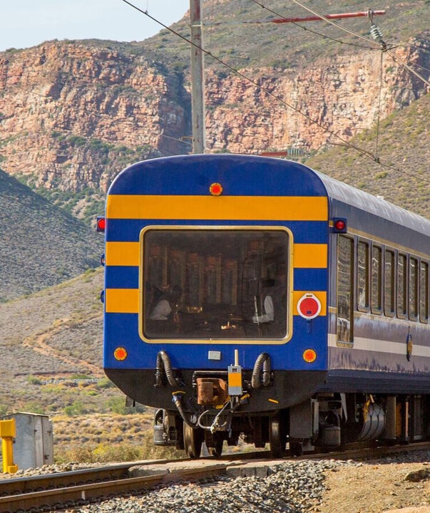 A blue and yellow train moves through a rocky mountain pass on luxury South Africa rail travel.