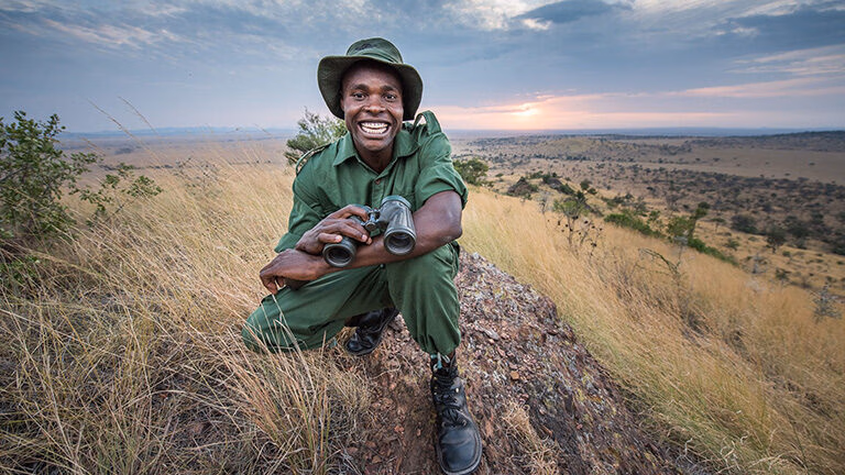 A smiling safari guide holding binoculars on a rocky outcrop during luxury Africa honeymoon tours at sunset.