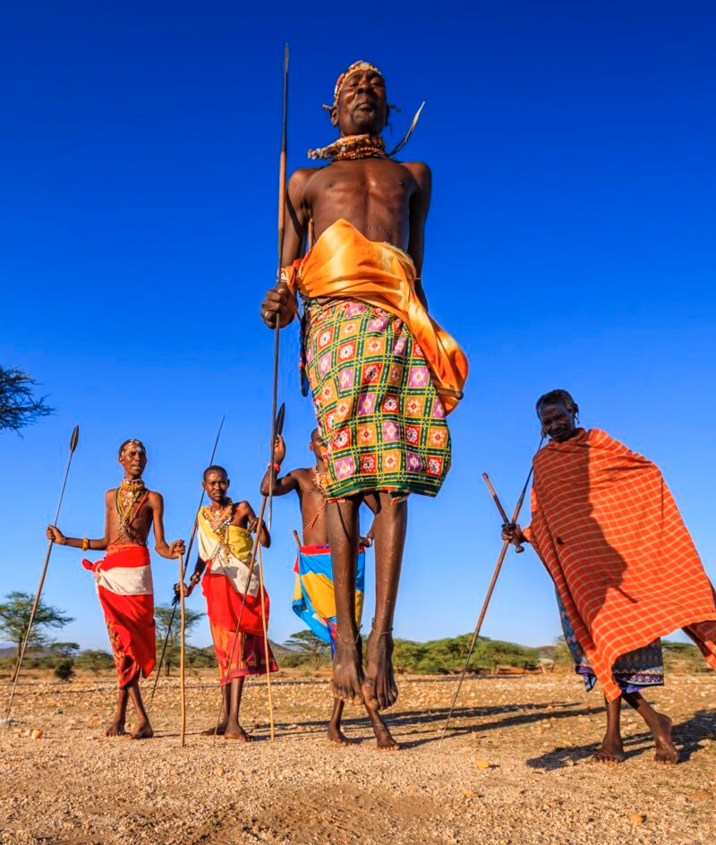 Maasai men in traditional red and patterned clothing performing a jumping dance on luxury Maasai Mara trips.