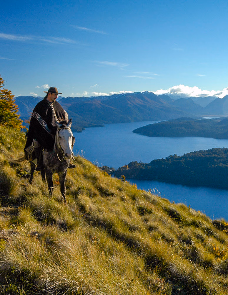 A person riding a horse on a high grassy path above a blue lake