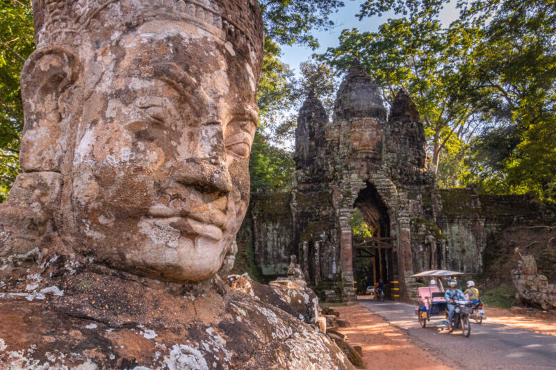 Close-up of a colossal, weathered stone face statue with the ornate, dark temple gate of Angkor Thom in the background.