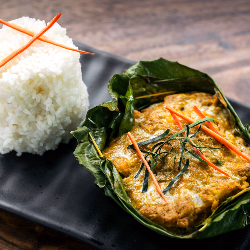 Traditional Cambodian Fish Amok, a yellow fish curry in a banana leaf, next to a tall, neat mound of white rice.