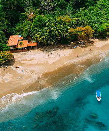 High-angle view of a sandy tropical beach, clear blue water with a small boat, and thick green forest.