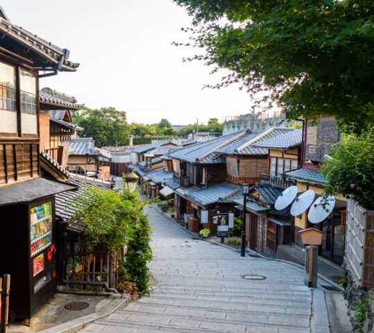 Sloping stone street in Japan with traditional wooden shops and green trees under a bright sky.