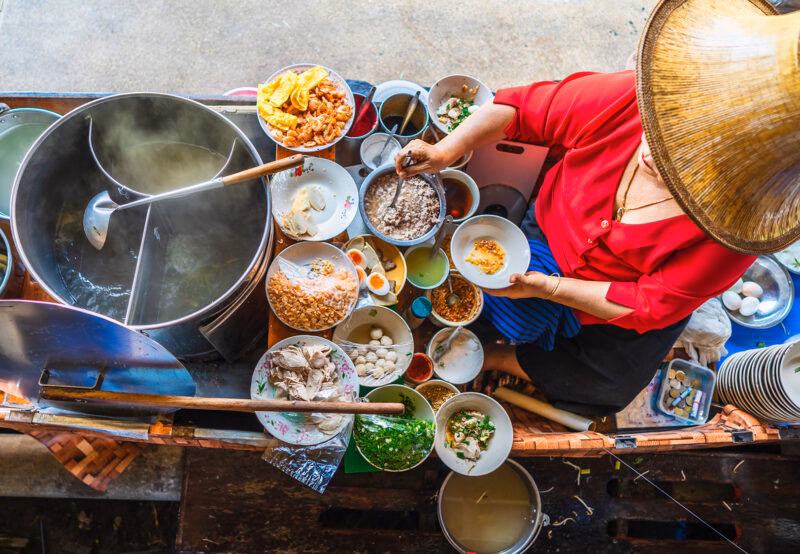 A street food vendor in a conical hat prepares bowls of noodles and various ingredients at a floating market. luxury food and wine tours.