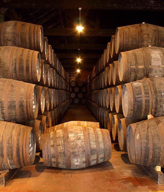 Rows of stacked wooden aging barrels in a large, dimly lit cellar or warehouse. luxury food and wine trips.