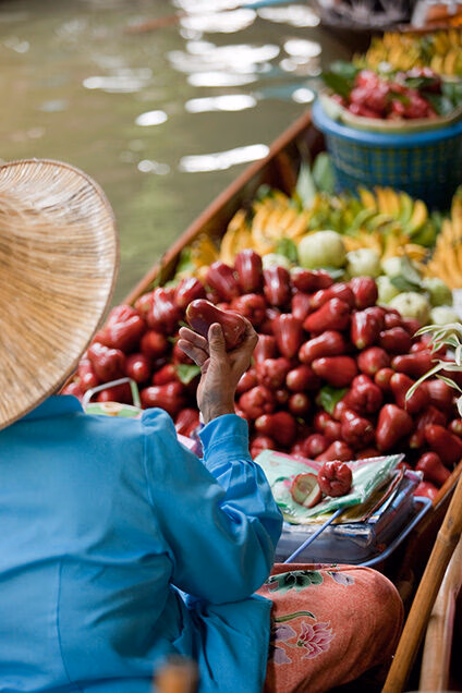 A vendor in a blue shirt holds a red fruit in a boat filled with tropical fruit. luxury food and wine tours