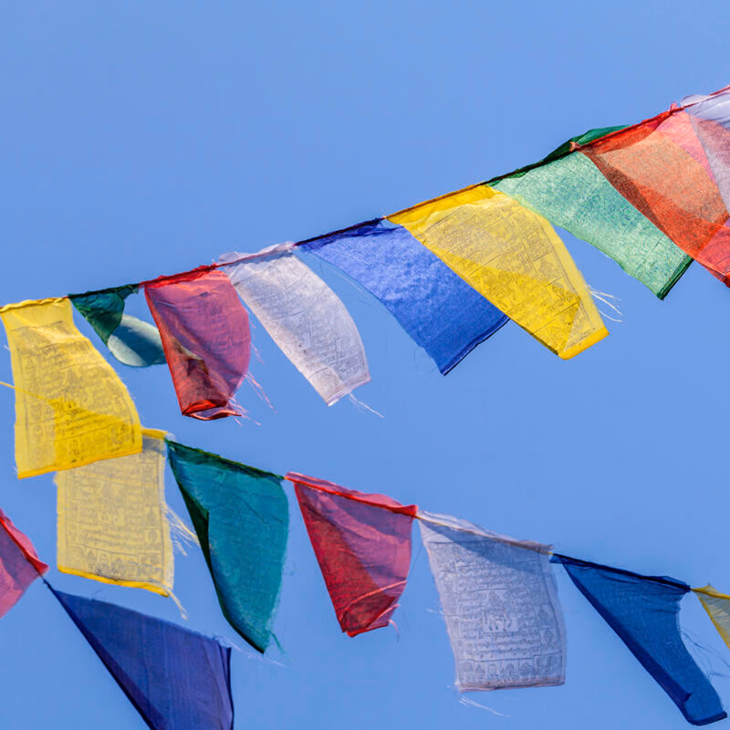 Multiple rows of colorful rectangular prayer flags strung across a vivid blue sky.