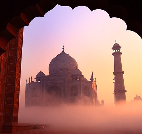 The Taj Mahal shrouded in pink morning fog viewed through an archway during luxury history tours.