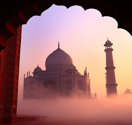 The Taj Mahal shrouded in pink morning fog viewed through an archway during luxury history tours.