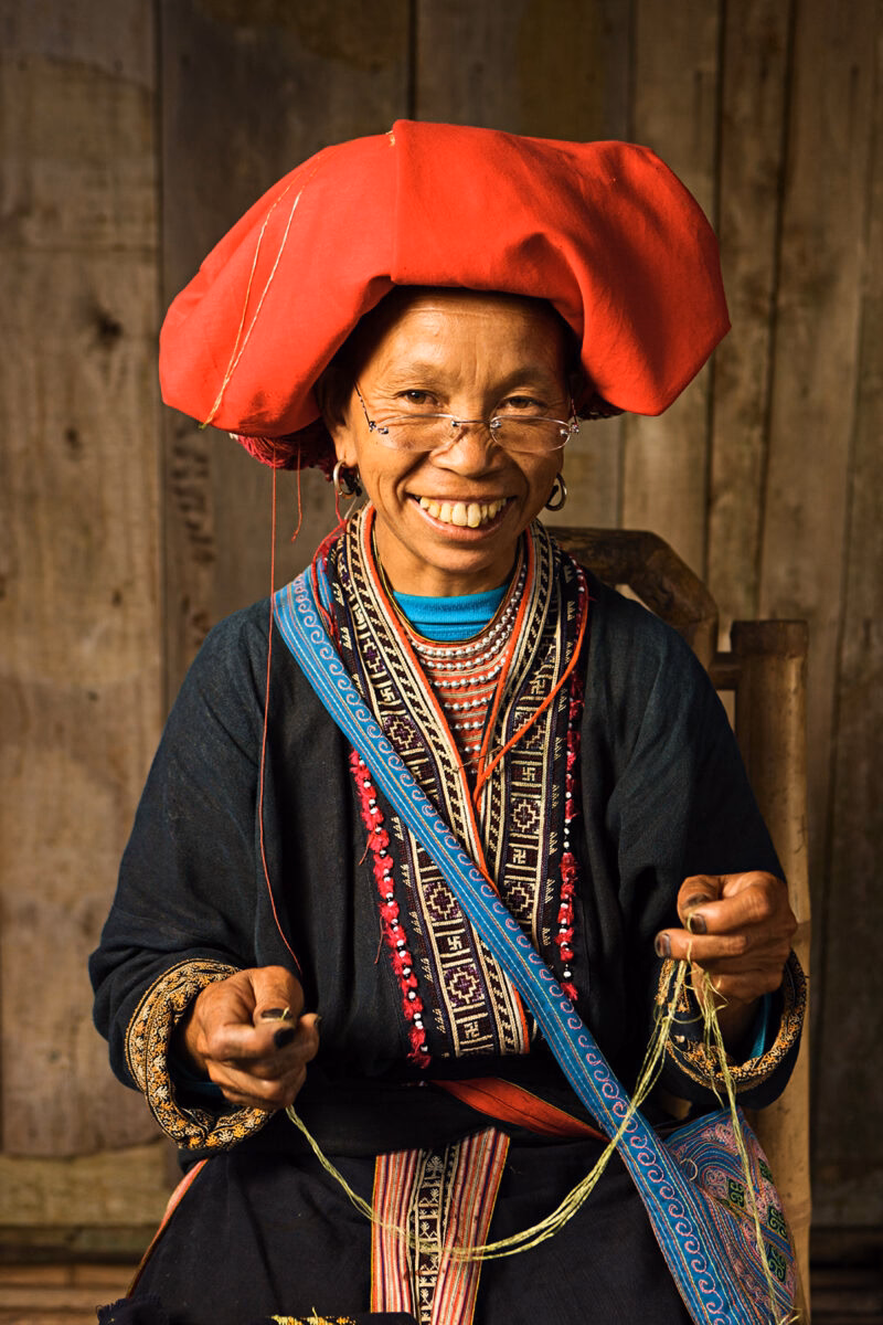 Woman in a large red headdress and traditional embroidered clothing sewing for luxury culture tours.