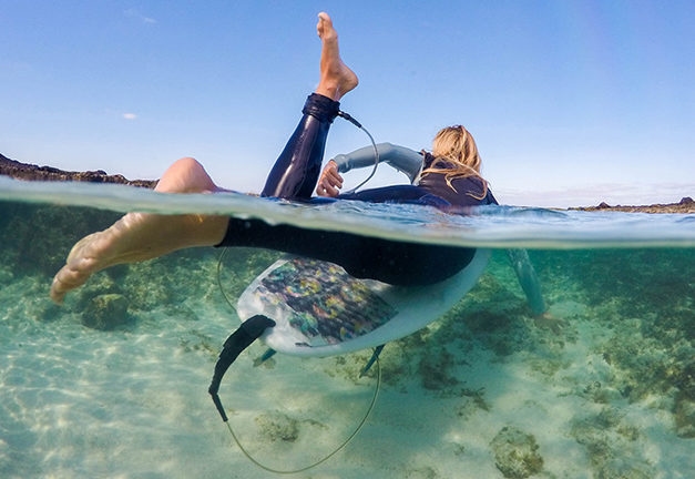 A surfer in a wetsuit resting on a surfboard in crystal clear water during luxury adventure trips.