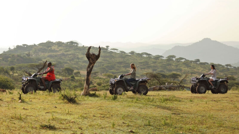 Three people riding ATVs in a line across a grassy plain with hills in the background on luxury African family safari tours.