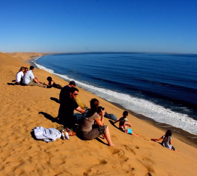 People sitting on a steep sand dune next to the blue ocean during luxury African family safari holidays.