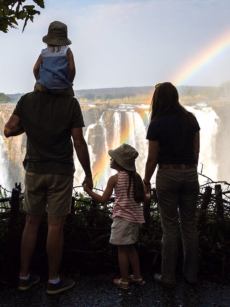 Rear view of a family looking at a large waterfall with a prominent rainbow during African family safaris.