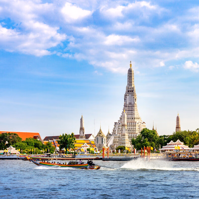A traditional long tail boat filled with people in front of Wat Arun Temple, a dramatic spire-shaped white and gold temple
